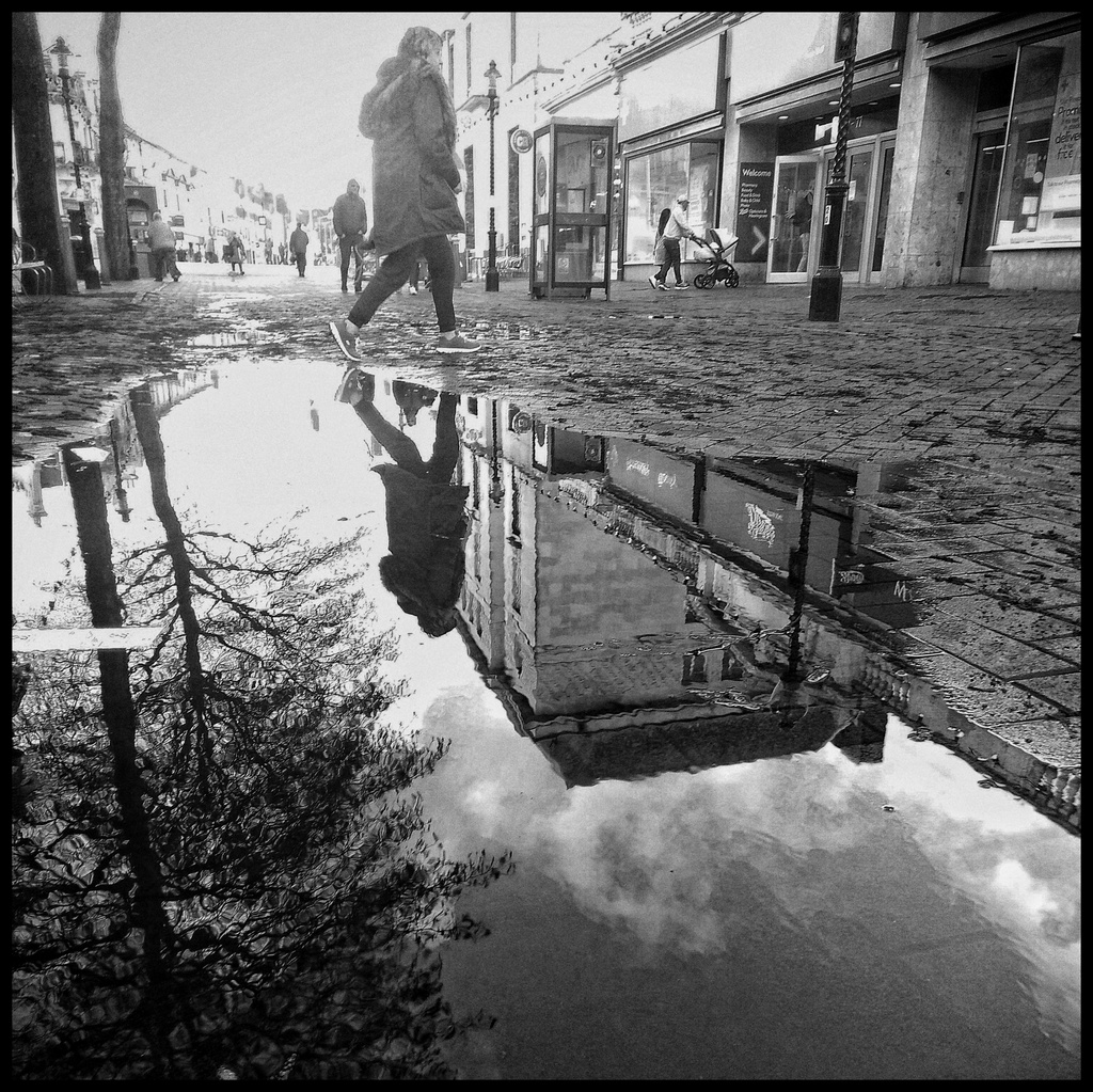 Black and white photo of a puddle reflecting buildings and a person walking across the street
