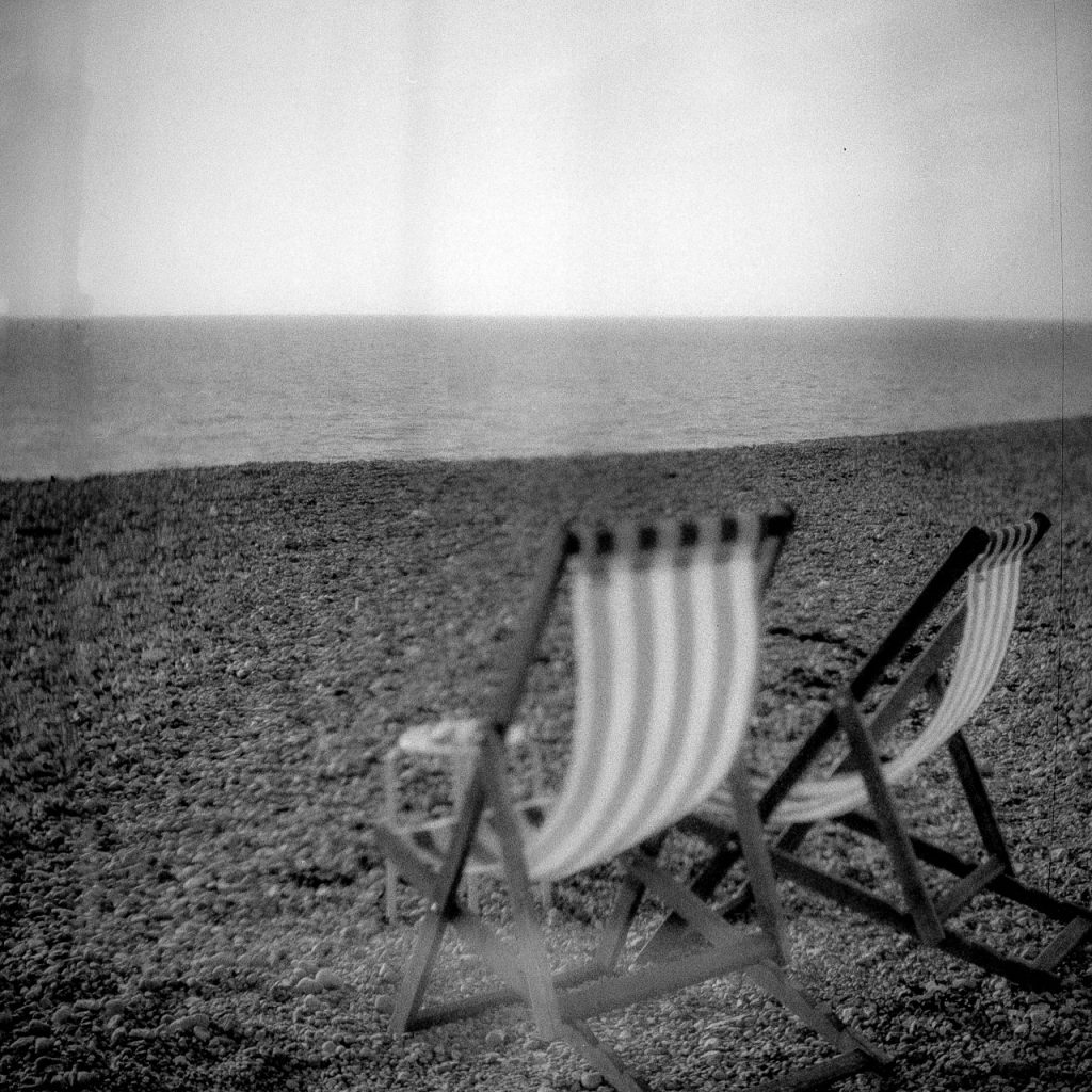 Black and white photo of two deckchairs sitting in front of the sea. The closer deckchair is blurred.