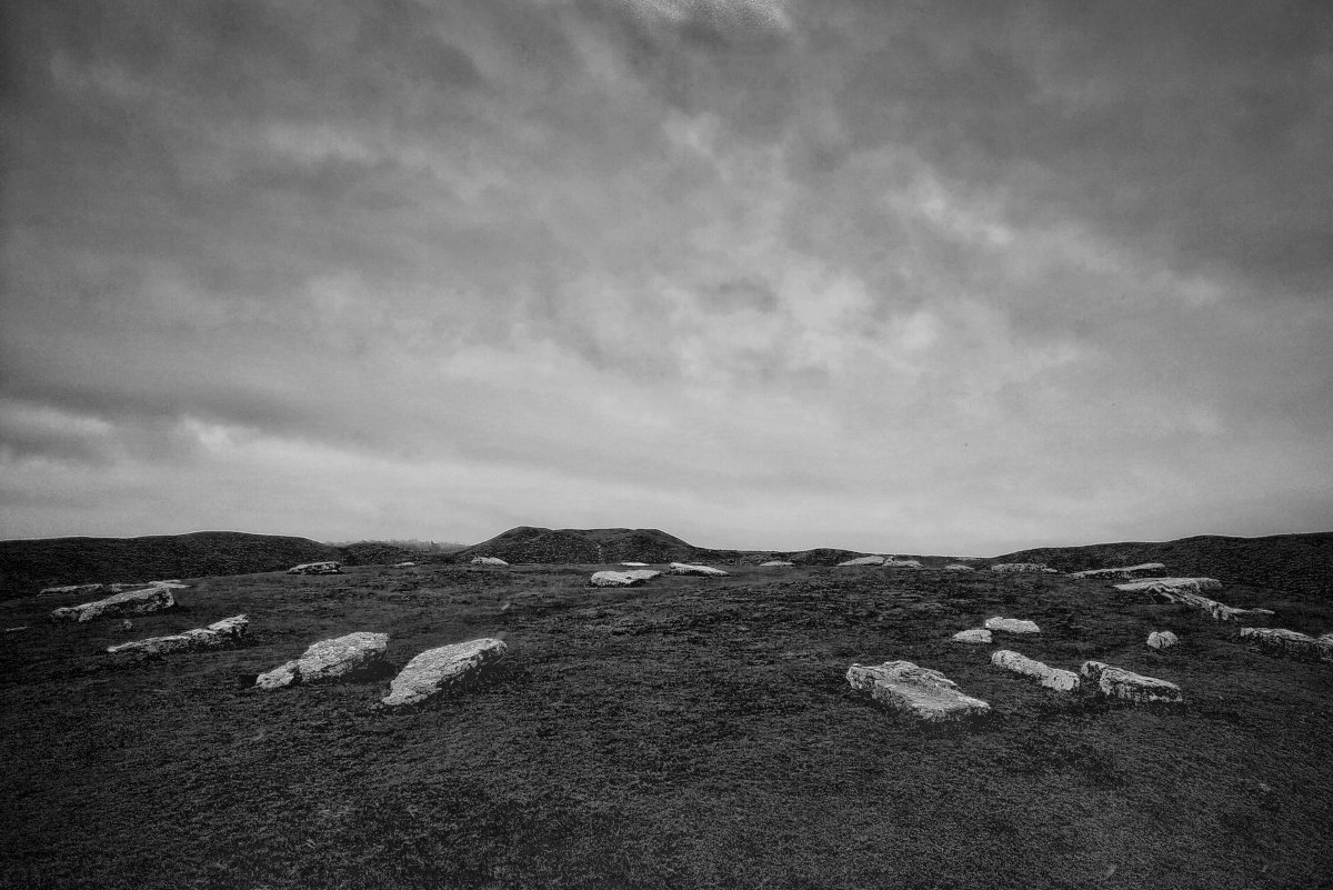 Black and white photo of dark ground under gloomy sky. A few large rocks are laid flat in a line along the ground, showing the edge of Arbor Low stone circle. A few more rocks are positioned in the middle of the ground.