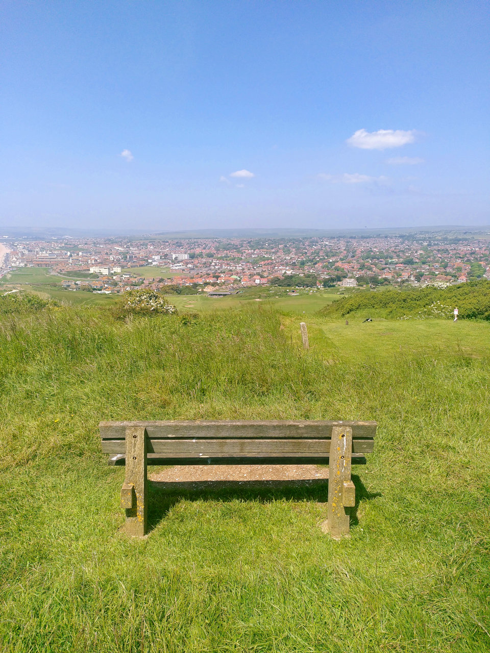 Photo of a bench on top of a grassy hill, overlooking a small town in the distance, under blue sky with a couple of fluffy clouds.