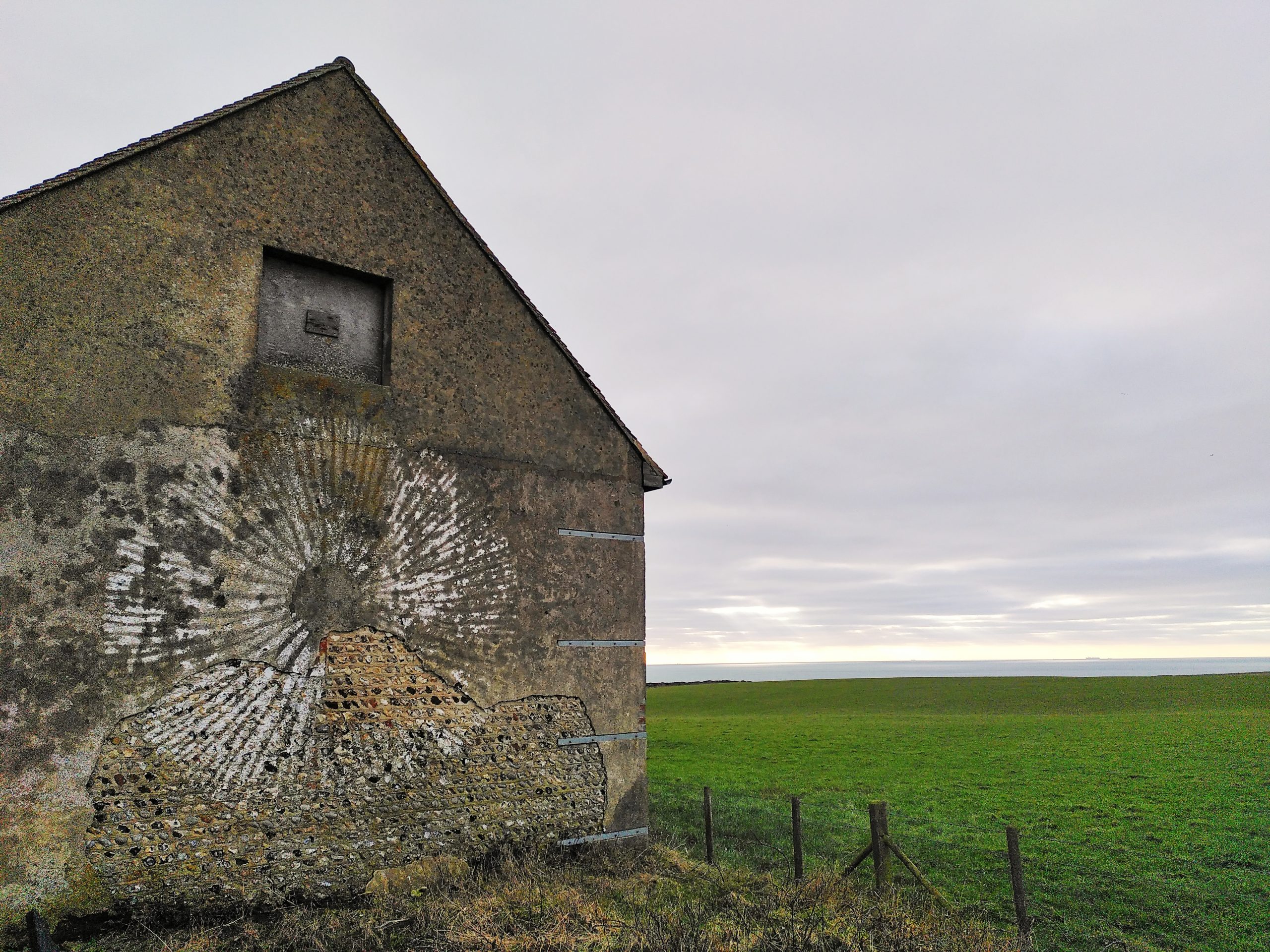 Photo of a large, old barn building beside a field. The barn has a blocked up window, and a white circular pattern painting on the side.